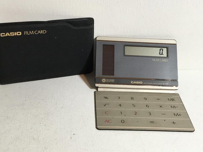May include: A vintage Casio Film Card calculator with a gray and silver design, featuring a numeric keypad and a digital display showing "0". The calculator is open, next to a black case with "CASIO FILM CARD" printed in gold.