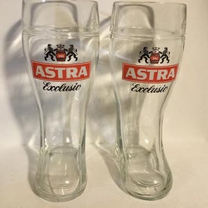 May include: Two clear glass beer boots with the "ASTRA Exclusiv" logo in red and white. The logo features a crest with two lions. The glasses are shaped like boots and are empty.