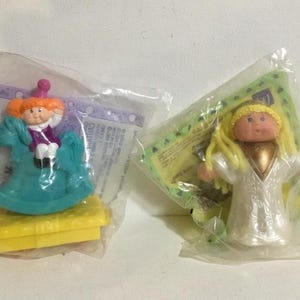 Puede incluir: Dos muñecas Cabbage Patch Kids en su embalaje original. Una muñeca está montada en un caballo de balancín y la otra muñeca lleva un vestido blanco con ribetes dorados.