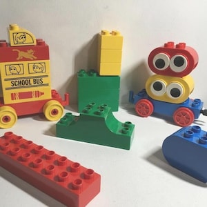 May include: A set of colourful Lego blocks, including a red school bus with yellow wheels, a green and yellow train, and a blue and yellow robot. The blocks are all made of plastic and are designed for children to build with.