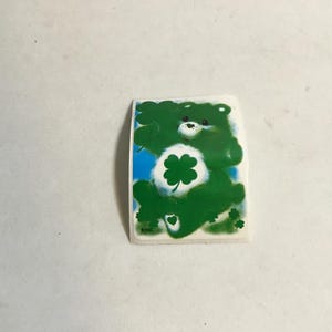 May include: Green Care Bear sticker with a four-leaf clover on its chest. The Care Bear is smiling and has a white face with black eyes.