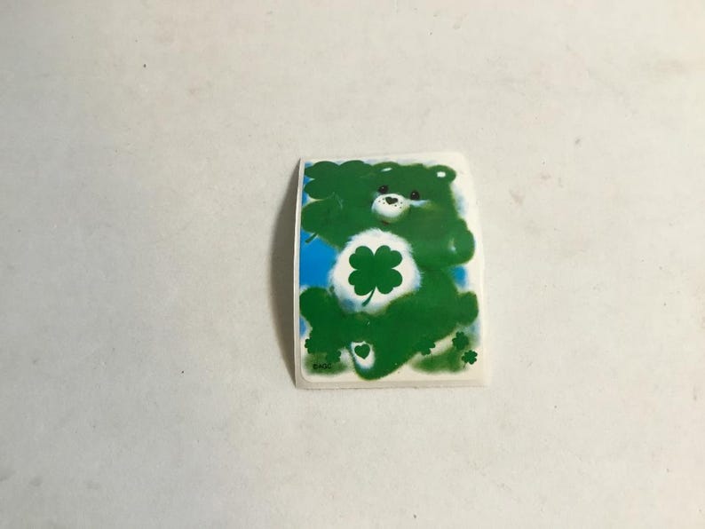 May include: Green Care Bear sticker with a four-leaf clover on its chest. The Care Bear is smiling and has a white face with black eyes.