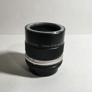 May include: Black teleconverter lens with silver markings. The lens has the text "Custom Tele-Converter" printed on the side.