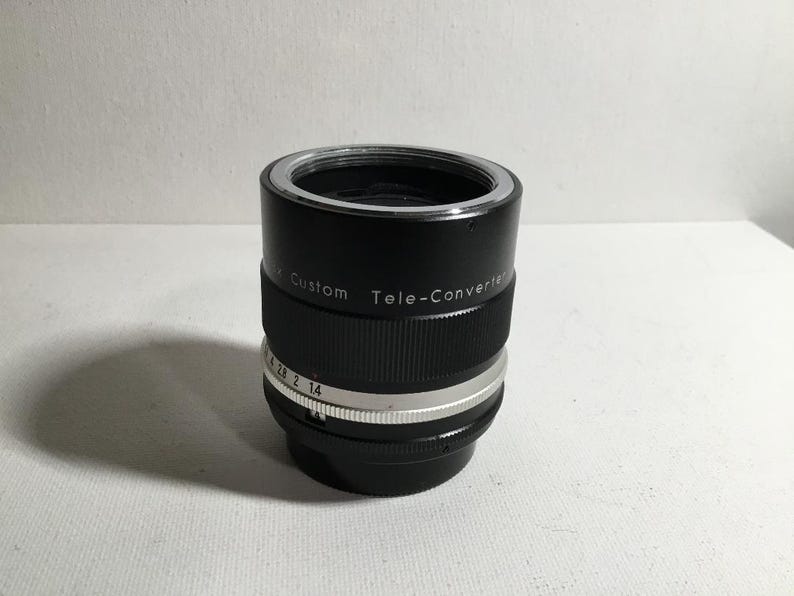 May include: Black teleconverter lens with silver markings. The lens has the text "Custom Tele-Converter" printed on the side.