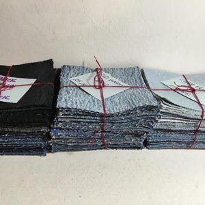May include: Three stacks of fabric squares in varying shades of blue and black, each tied with red twine. The stacks are labeled with handwritten notes indicating the color and quantity, such as "Black 4x4 25 Pack".
