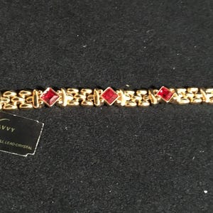 May include: A gold-tone link bracelet featuring three square-cut red crystals. Attached to the bracelet is a black rectangular tag with the text "SAVVY" in gold lettering and "SWAROVSKI FULL LEAD CRYSTAL" printed in smaller white text below.