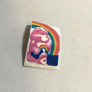 May include: A pink Care Bear with a rainbow and a heart on its chest is sitting on a blue box. The Care Bear is holding a rainbow in its paws. The sticker is on a white background.