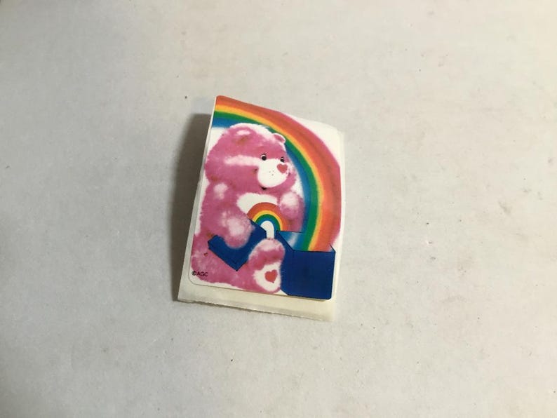 May include: A pink Care Bear with a rainbow and a heart on its chest is sitting on a blue box. The Care Bear is holding a rainbow in its paws. The sticker is on a white background.