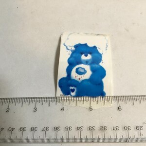 May include: A blue Care Bear sticker with a cloud on its belly and a white cloud above its head. The Care Bear is holding a heart with a cloud inside. The sticker is about 2 inches tall.