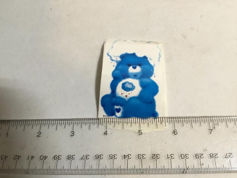 May include: A blue Care Bear sticker with a cloud on its belly and a white cloud above its head. The Care Bear is holding a heart with a cloud inside. The sticker is about 2 inches tall.