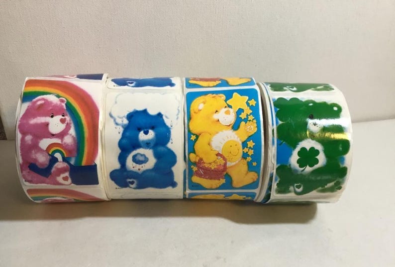 May include: Five rolls of Care Bear stickers featuring different Care Bears. The stickers are on a white background and feature a variety of colors, including pink, blue, yellow, and green.