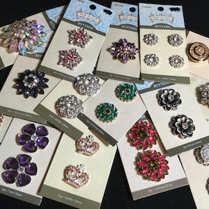 May include: A collection of Sew-Ology brand buttons displayed on individual cardboard backing cards. The assortment includes various decorative shapes such as flowers, crowns, and geometric patterns featuring clear, pink, purple, green, and gold-toned rhinestones and metallic finishes.