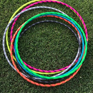 Op de afbeelding: Een set kleurrijke plastic hoepels, op elkaar gestapeld, op een groene grasmat. De hoepels zijn in verschillende kleuren, waaronder neon groen, oranje, turquoise, grijs en roze.