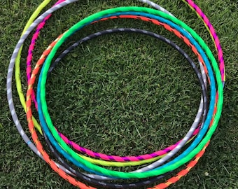 May include: A set of colorful plastic hula hoops, stacked on top of each other, on a green grassy surface. The hoops are various colors, including neon green, orange, teal, gray, and pink.