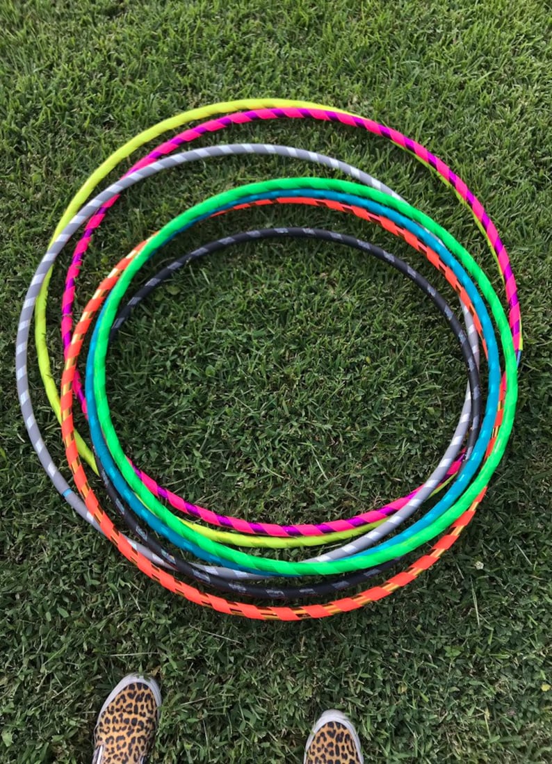 Op de afbeelding: Een set kleurrijke plastic hoepels, op elkaar gestapeld, op een groene grasmat. De hoepels zijn in verschillende kleuren, waaronder neon groen, oranje, turquoise, grijs en roze.