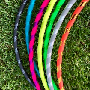 Op de afbeelding: Een set van acht kleurrijke hoelahoepringen met verschillende gestreepte patronen. De ringen zijn gemaakt van plastic en hebben een glanzende afwerking. De ringen zijn in een halve cirkel op een groene grasmat gerangschikt.