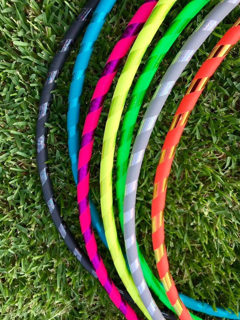 Op de afbeelding: Een set van acht kleurrijke hoelahoepringen met verschillende gestreepte patronen. De ringen zijn gemaakt van plastic en hebben een glanzende afwerking. De ringen zijn in een halve cirkel op een groene grasmat gerangschikt.