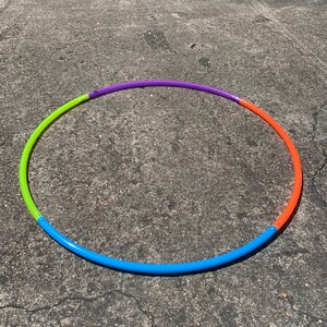 May include: A colorful hula hoop with segments of blue, green, purple, and orange. The hoop is lying on a gray concrete surface, suggesting outdoor use. The segments are connected with small metal connectors.