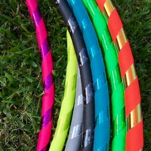Op de afbeelding: Een set kleurrijke hoelahoepringen met verschillende patronen en kleuren, waaronder roze, groen, blauw, zwart en goud. De ringen zijn in een halve cirkel gerangschikt, met de roze ring op de voorgrond.