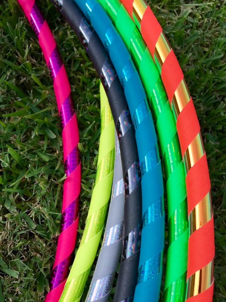 Op de afbeelding: Een set kleurrijke hoelahoepringen met verschillende patronen en kleuren, waaronder roze, groen, blauw, zwart en goud. De ringen zijn in een halve cirkel gerangschikt, met de roze ring op de voorgrond.
