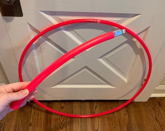 May include: A bright pink exercise hoop, also known as a hula hoop, is lying on a wooden floor. The hoop is partially disassembled, with one section of the hoop lying on the floor and the other section being held by a hand.