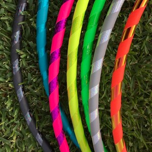 Op de afbeelding: Een set van zes kleurrijke hoelahoepringen met verschillende patronen en kleuren, waaronder zwart, teal, roze, geel, grijs en oranje. De ringen zijn in een waaier vorm gerangschikt, met de zwarte ring onderaan en de oranje ring bovenaan.