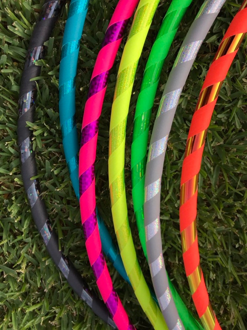 Op de afbeelding: Een set van zes kleurrijke hoelahoepringen met verschillende patronen en kleuren, waaronder zwart, teal, roze, geel, grijs en oranje. De ringen zijn in een waaier vorm gerangschikt, met de zwarte ring onderaan en de oranje ring bovenaan.