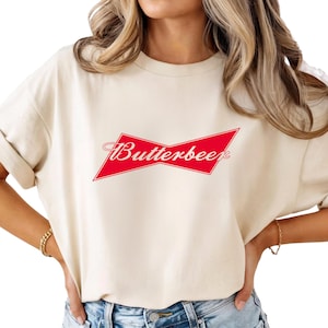 May include: Cream-colored t-shirt featuring a red and white graphic that reads "Butterbeer." The design is a parody of a well-known beer logo. The shirt has rolled-up sleeves and a relaxed fit.