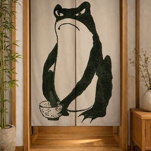 May include: A beige fabric noren curtain featuring a large, dark green, stylized illustration of a frog with a grumpy expression, holding a small speckled bowl. The curtain is split down the center and hangs within a wooden door frame.