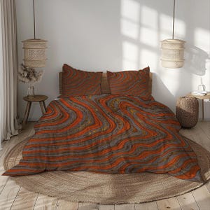 May include: A bed with a duvet cover and two pillowcases featuring a swirling pattern in shades of orange, gray, and gold. The bedding is on a wooden bed frame, with a round rug beneath. The room has a neutral color palette with woven pendant lights.