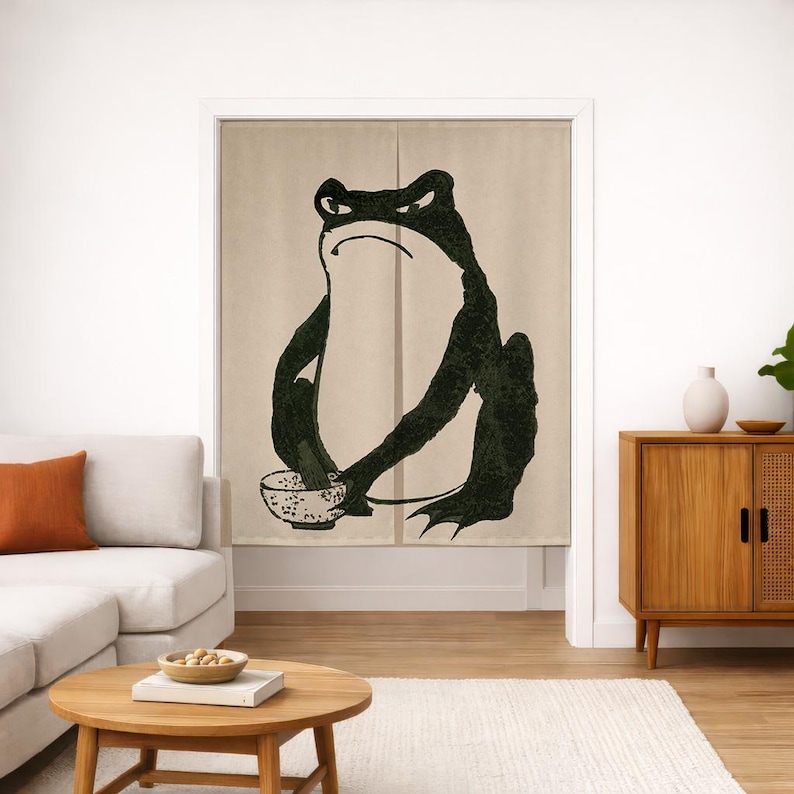 May include: A beige fabric door curtain featuring a large, dark green, stylized illustration of a frog sitting upright and holding a small, speckled bowl. The curtain is split down the center and hangs vertically within a doorway frame.