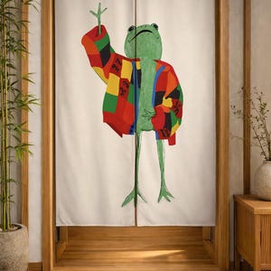 Pode incluir: Uma cortina noren de tecido bege com uma ilustração central de um sapo verde vestindo um cardigã de patchwork colorido com quadrados vermelhos, amarelos, verdes e pretos. O sapo é retratado com um braço levantado e duas pernas finas.