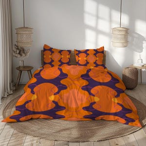May include: A double bed with an orange, purple, and yellow patterned duvet cover and two matching pillowcases. The design features large, abstract shapes. The bed is made up and ready for use. The room has a neutral colour scheme.