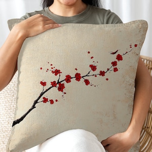Op de afbeelding: Een beige kussen met een rood en zwart bloemenontwerp. Het ontwerp toont een gestileerde tak met rode bloemen en een kleine vogel.