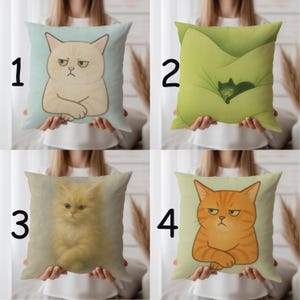 May include: Four decorative throw pillows with cat illustrations. The first pillow has a beige cat with a grumpy expression on a light blue background. The second pillow has a green cat on a green background. The third pillow has a fluffy yellow cat. The fourth pillow has an orange cat with a grumpy expression.