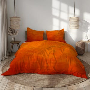 Op de afbeelding: Een oranje en rode ombre dekbedovertrekset met twee bijpassende kussenslopen. Het beddengoed wordt weergegeven op een houten bedframe, met een neutraal gekleurd tapijt en decoratieve hanglampen op de achtergrond. Het algemene thema is woondecoratie.