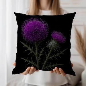 Op de afbeelding: Zwart vierkant kussen met een gedetailleerde illustratie van paarse distelbloemen met stekelige groene bladeren en stengels. De bloemen staan in volle bloei, tegen een effen zwarte achtergrond. Het kussen wordt door een persoon vastgehouden.