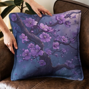 Puede incluir: Cojín cuadrado con fondo azul oscuro y diseño floral morado. El cojín presenta un árbol con flores moradas. El cojín está colocado sobre un sofá de cuero marrón.