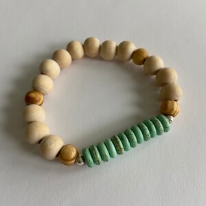 Könnte beinhalten: Ein Holzperlenarmband mit einem türkisfarbenen Akzent. Das Armband besteht aus natürlichen Holzperlen in Braun- und Weißtönen. Die türkisfarbenen Perlen sind in einer Reihe in der Mitte des Armbands angeordnet.