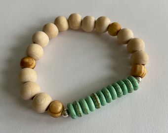 Può includere: Un braccialetto di perline di legno con un accento turchese. Il braccialetto è fatto di perline di legno naturale in tonalità di marrone e bianco. Le perline turchesi sono disposte in una fila al centro del braccialetto.