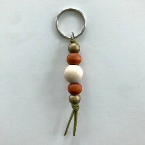 May include: A wooden bead keychain with a silver key ring. The keychain has three beads: one white, one orange, and one gold. The keychain also has a green leather tassel.