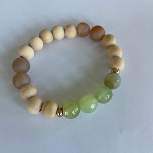 Op de afbeelding: Een armband gemaakt van lichtbruine houten kralen en groene jade kralen. De armband heeft een verloop van bruine kralen, beginnend met een donkerder bruin en vervagend naar een lichter bruin.