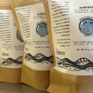 May include: Four brown paper bags with white labels. The labels have black and white illustrations of bubbles and text that reads "Handmade Stinky Vest Vegan Soap Company", "Fizzy Bubble Bath Powder Juniper Alice", "Net Wt 4 oz (114g)", "Stinky Vest Vegan Soap Co", "Fizzy Bubble Bath Powder Bright 'n Early", and "All products are poured by hand in Boulder, CO".