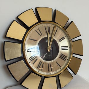 Puede incluir: Un reloj de pared de cuarzo dorado y negro con un diseño de rayos de sol. La esfera del reloj tiene números romanos y agujas doradas. El reloj es redondo con un borde negro y detalles dorados.