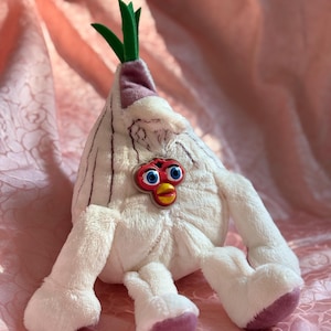 May include: A plush toy shaped like an onion with a Furby face. The toy is primarily white with purple accents on the feet and top. Green fabric forms the onion's sprouts. The Furby face has blue eyes and a red beak.