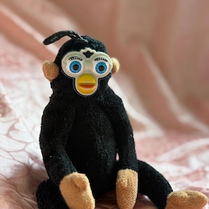 Puede incluir: Un juguete Furby negro vintage con detalles en color beige. El Furby tiene ojos azules grandes, un pico amarillo y orejas y pies beige. El juguete está sobre una superficie rosa.