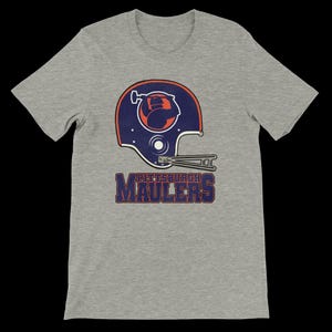 Puede incluir: Camiseta gris con un diseño de casco de fútbol de estilo vintage en azul marino, naranja y blanco. El texto "PITTSBURGH MAULERS" está impreso debajo en azul marino y naranja.
