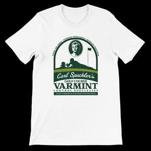 May include: White t-shirt with a green graphic featuring the text "Carl Spackler's Golf Course Varmint Control Specialist." The design includes an illustration of a person, a golf course, and the words "Knows the Enemy, Superior Intelligence, Superior Firepower."