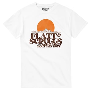 Può includere: T-shirt bianca con un design retrò. Il design presenta la scritta "Flatt & Scruggs with The Foggy Mountain Boys" in marrone, un sole arancione e montagne. La maglietta ha un girocollo.