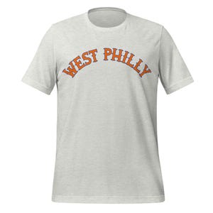 Könnte beinhalten: Hellgraues T-Shirt mit dem orangefarbenen Schriftzug "WEST PHILLY" in einem Bogen über der Brust. Das T-Shirt hat kurze Ärmel und einen Rundhalsausschnitt. Der Text ist in einer kräftigen, stilisierten Schriftart gehalten.